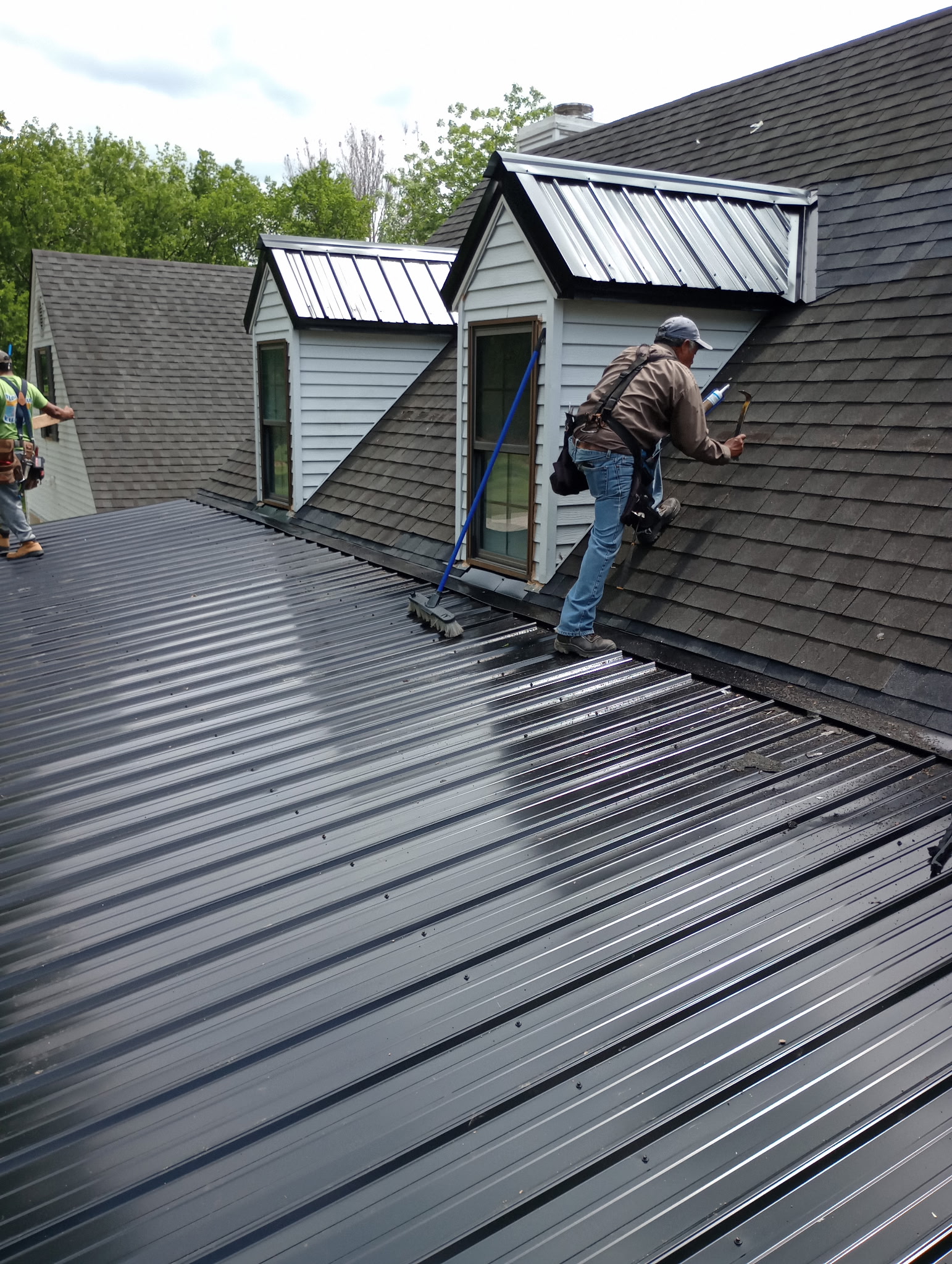 Standing-seam metal roof being installed beside asphalt shingles and dormers